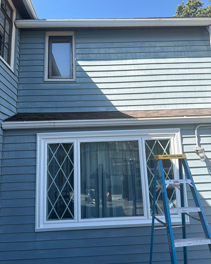Blue siding in Drexel Hill, PA partway through oxidation removal, the upper story still chalky and the lower story back to its deep blue, a ladder at the window