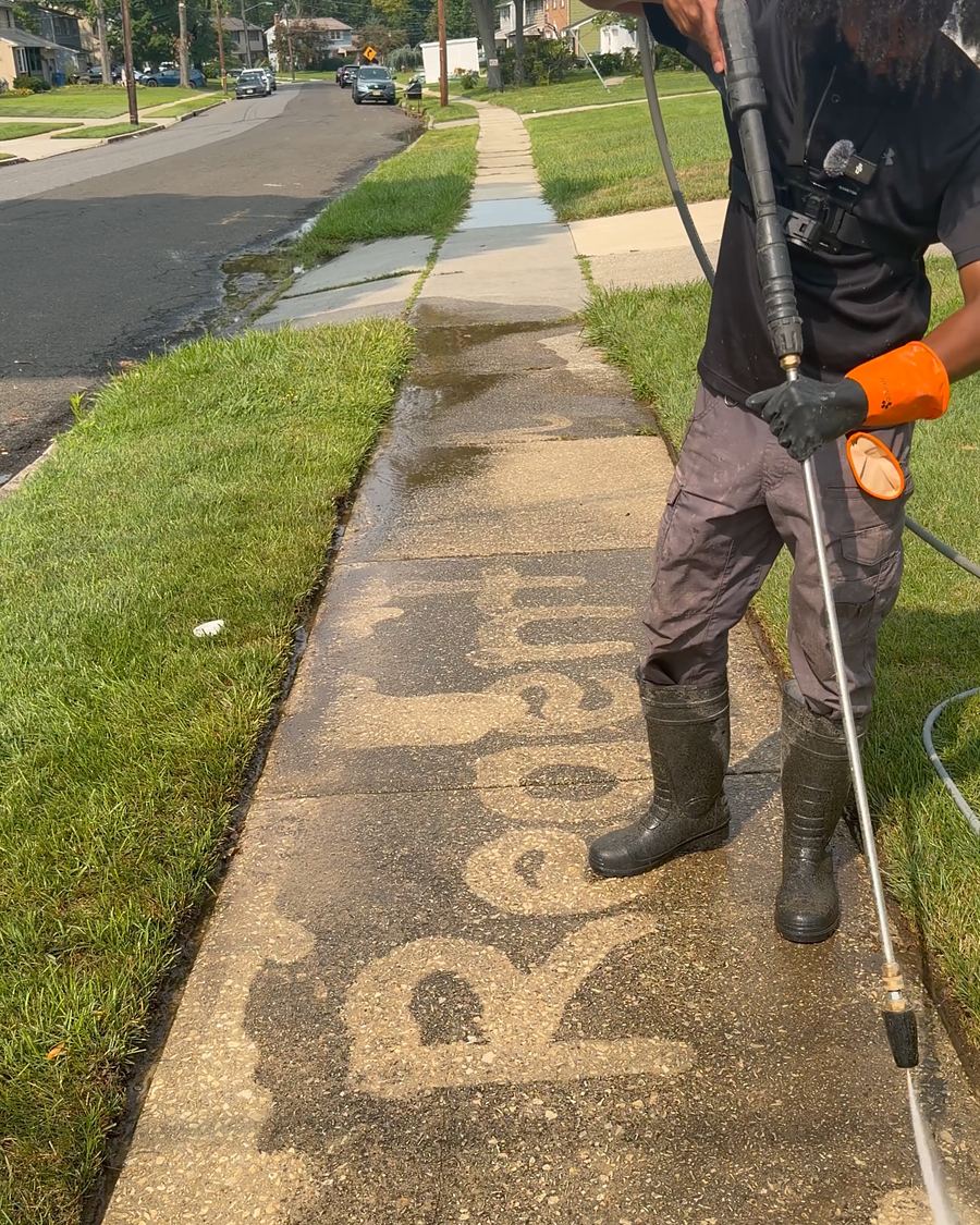A Regent Wash technician pressure washing a concrete sidewalk, the cleaned section visible behind them