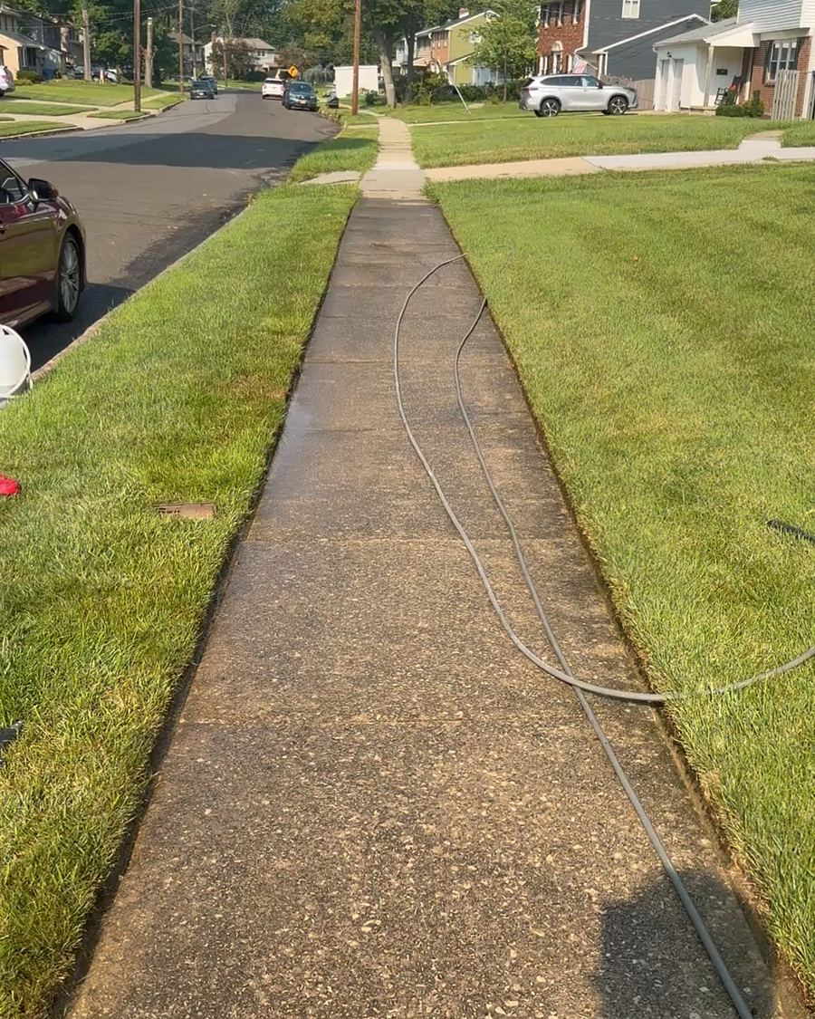 A long public concrete sidewalk before pressure washing, the slabs dark with grime along the whole block